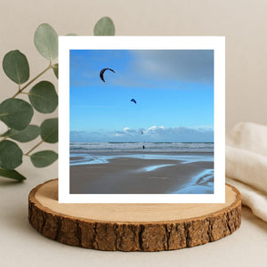 Beach scene with kites in the sky and kite surfers on the front of a greetings card displayed on a wooden stand.