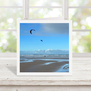 Photograph of a beach scene with kitesurfers against a blue sky on the front of a greetings card with white border.