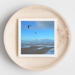 Photograph on a greetings card of a beach scene with kites surfers and kites flying against a blue sky.