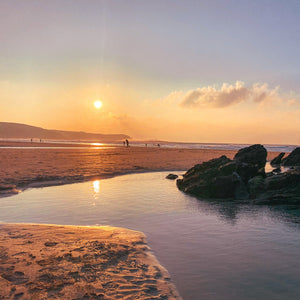 Sunset at a beach with people walking along the shore and large rocks  and rockpools in the foreground.