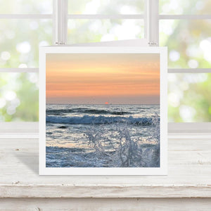 Greetings card of a sunset over the ocean with sea splash, displayed on a wooden surface with a blurred window background.