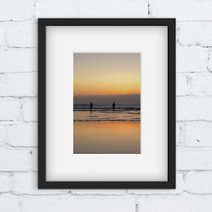 Framed photograph of two people going for a swim in the sea at sunset. Framed against a white brick wall