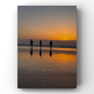 Three people standing on a beach at sunset, reflected in the wet sand.