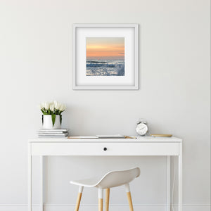 Framed sunset artwork above a white desk with a chair, clock, and flowers.