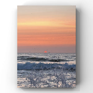 Pale orange and pink sunset with orange sun on the horizon. There is a splash of ocean wave in the foreground