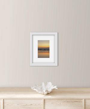 Framed artwork of two people going for a sea swim at sunset on a wall above a wooden surface with a decorative bowl.