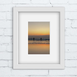 Framed artwork of a sunset over water with two silhouettes, on a white brick wall.