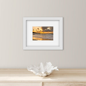Framed artwork of a sunset over water on a wall above a wooden surface with a decorative shell.