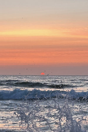 Pale orange and pink sunset with orange sun on the horizon. There is a splash of ocean wave in the foreground
