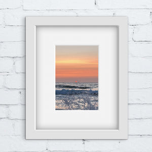 Pale orange pink sunset with ocean splash in a white frame against a white brick wall