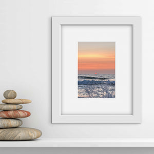 Pale orange and pink sunset photo with ocean water splash in a white frame and pile of flat pebbles