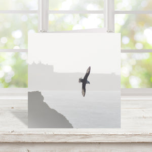 A square art print featuring a black bird in flight over a misty gray sea and coastline, displayed on a white wooden surface in front of a softly blurred window.