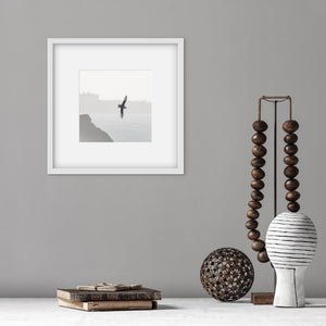 square white frame with an image of a soaring bird above sea cliffs. The picture s on a grey wall with wooden trinkets on a shelf below