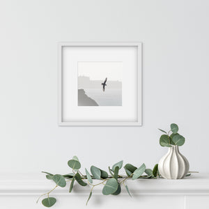 A framed black and white art print depicting a bird flying over water, styled on a white mantelpiece above a white vase with green eucalyptus branches.