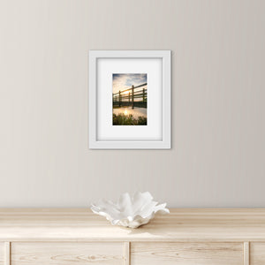 Framed photograph of a bridge over water  and sunset, on a wall above a wooden cabinet.