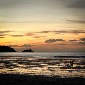 Two people walking on a beach at sunset with a silhouetted headland in the background
