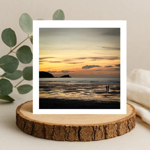 Sunset over a beach with two people standing on the shore, greetings card framed by a white border and sitting on a wooden coaster.