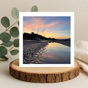 Greetings card of a sunset reflected  in water . Displayed on a wooden stand with a plant and blanket in the background.