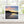 Square greetings card with photograph of a sunset over a beach with boats. Displayed on a wooden surface.