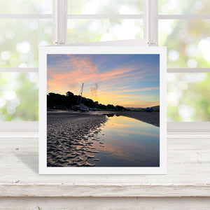 Square greetings card with photograph of a sunset over a beach with boats. Displayed on a wooden surface.