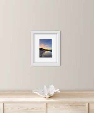 Framed photograph of a pastel sunset over water on a wall above a wooden cabinet with a decorative bowl.