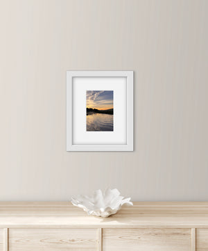 Framed photograph of a sunrise over water on a wall above a wooden cabinet with a decorative bowl.