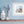 Sea pink flowers and bee in a white frame against a blue wall. There is a wooden shelf below with trinkets