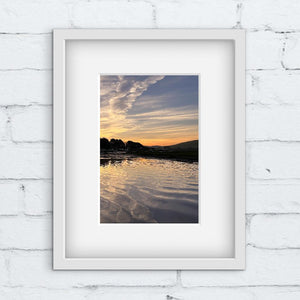 Framed photograph of a sunrise over water on a white brick wall.