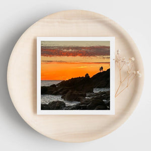 Greetings card of an sunset over a rocky landscape with two fishermen. Displayed on a beige plate.