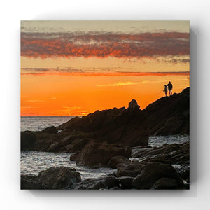 Two people fishing standing on a rocky outcrop at sunset with an orange sky.