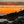Two people standing on rocks fishing at sunset with a vibrant orange sky.