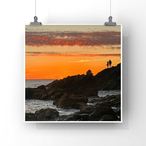 Two people standing on rocks fishing at sunset with a vibrant orange sky.