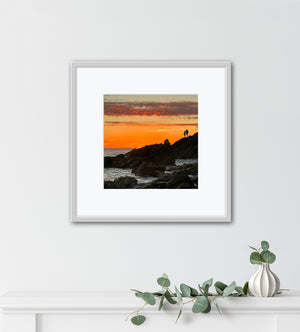 Framed artwork of a sunset over a rocky landscape with two people on a cliff fishing, displayed on a white wall above a mantel with decorative elements.