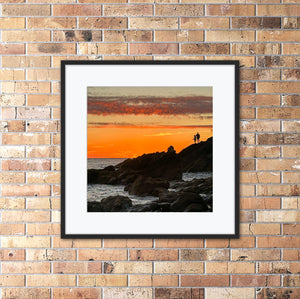 Framed artwork of a sunset over water with two figures on a rock fishing, mounted on a brick wall.