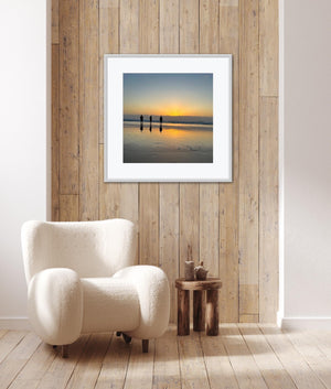 White armchair in a room with wooden walls and a framed picture of a sunset and people silhouetted over water.