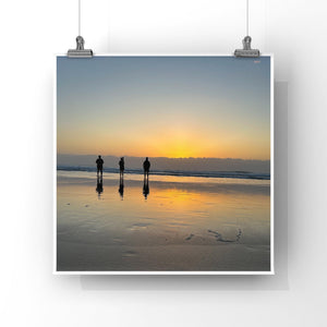 Three people standing on a beach at sunset, with their reflections visible in the wet sand.