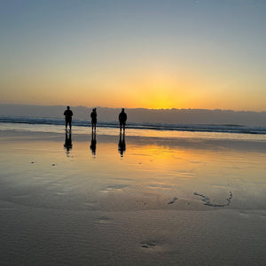 Three people standing on a beach at sunset, with their reflections visible in the wet sand.