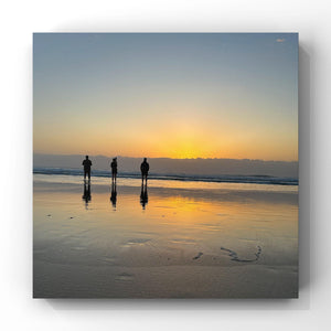 Three people standing on a beach at sunset, with their reflections visible in the wet sand.
