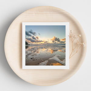White edged beach sunset greetings card on a round beige surface with dried flowers.