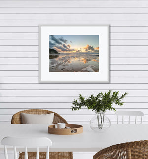 Framed beachscape sunset print reflected on wet sand, on a white wall above a dining table with chairs.