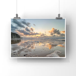 Beach scene with sunset, clouds reflecting on water, and cliffs. on print hanging from 2 wires