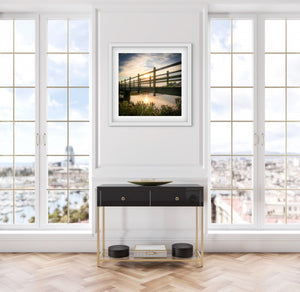 Modern console table with a framed picture above of a sunset behind a wooden bridge in a room with large windows.