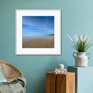 Framed beach print on a teal wall with a wooden side table and plant.