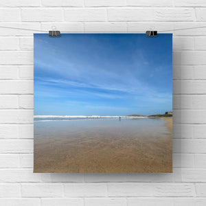 Print of a beach scene with blue sky and ocean on a white brick wall.