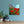 Unframed print of red poppies on a blue wall with two potted plants and a small sculpture on a wooden surface.