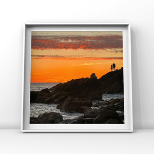 Framed artwork of a sunset over rocks with two silhouettes fishing, on a white wall.